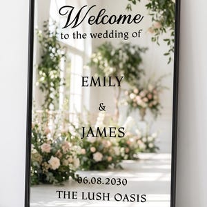 May include: A rectangular mirror with a thin black frame featuring black text that reads Welcome to the wedding of EMILY & JAMES 06.08.2030 THE LUSH OASIS. The mirror reflects a bright room with floral arrangements and greenery.