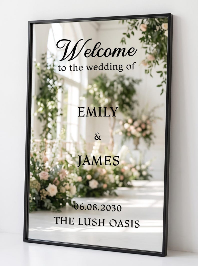 May include: A rectangular mirror with a thin black frame featuring black text that reads Welcome to the wedding of EMILY & JAMES 06.08.2030 THE LUSH OASIS. The mirror reflects a bright room with floral arrangements and greenery.