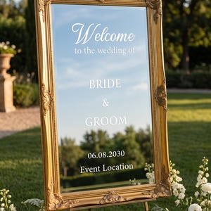 May include: A rectangular mirror with an ornate gold frame featuring white text that reads Welcome to the wedding of BRIDE & GROOM 06.08.2030 Event Location. The mirror is mounted on a stand and reflects the surrounding outdoor greenery.