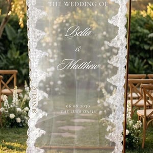 Pu&ograve; includere: Un cartello di benvenuto per matrimoni rettangolare con un pannello in tessuto bianco trasparente drappeggiato su una cornice in metallo dorato. Il tessuto &egrave; bordato da un intricato pizzo bianco e riporta il testo: welcome to THE WEDDING OF Bella & Matthew 06.08.2030 THE LUSH OASIS.