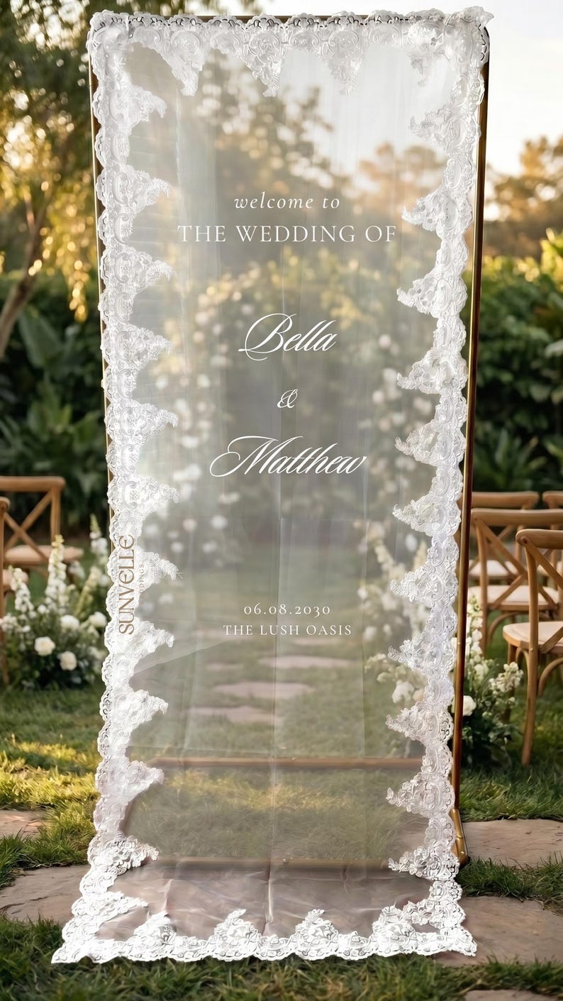 Pu&ograve; includere: Un cartello di benvenuto per matrimoni rettangolare con un pannello in tessuto bianco trasparente drappeggiato su una cornice in metallo dorato. Il tessuto &egrave; bordato da un intricato pizzo bianco e riporta il testo: welcome to THE WEDDING OF Bella & Matthew 06.08.2030 THE LUSH OASIS.