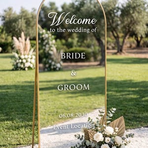 May include: A tall, arched, clear acrylic wedding welcome sign with a gold metal frame. The sign features white text reading Welcome to the wedding of BRIDE & GROOM 06.08.2030 Event Location. A floral arrangement with white roses sits at the base.