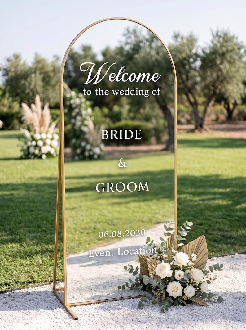 May include: A tall, arched, clear acrylic wedding welcome sign with a gold metal frame. The sign features white text reading Welcome to the wedding of BRIDE & GROOM 06.08.2030 Event Location. A floral arrangement with white roses sits at the base.