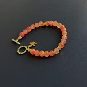 May include: A beaded bracelet featuring translucent orange round beads interspersed with small gold-toned square spacers. The bracelet includes a gold-toned toggle clasp and a small gold-toned four-leaf clover charm attached to the circular ring.