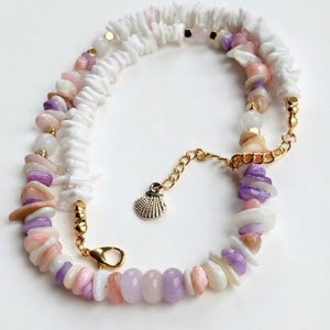 May include: A beaded necklace featuring a mix of white, lavender, and soft pink chip-style beads interspersed with small gold-toned metal spacers. The necklace includes a gold-toned lobster clasp and an extender chain with a small seashell charm.