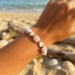 May include: A beaded bracelet featuring light pink stone chips, white heart-shaped beads, and small gold-toned metallic spheres. The bracelet includes a gold-toned butterfly charm and a short dangling chain extension with a single pink bead at the end.
