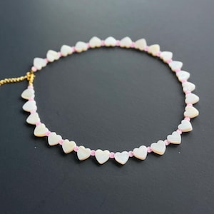Puede incluir: Un collar que presenta un patrón repetitivo de pequeñas cuentas blancas en forma de corazón alternadas con diminutas cuentas redondas de color rosa claro. El collar incluye una cadena extensora de metal dorado con un cierre de mosquetón.