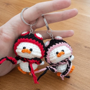 May include: Two handmade crochet penguin keychains. Each penguin has a white body, orange feet, and a black and red or black and pink hat and scarf. The keychains are held by a person.