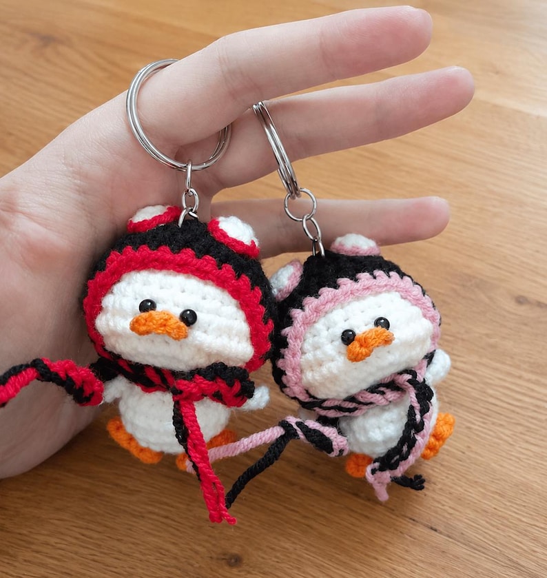 May include: Two handmade crochet penguin keychains. Each penguin has a white body, orange feet, and a black and red or black and pink hat and scarf. The keychains are held by a person.