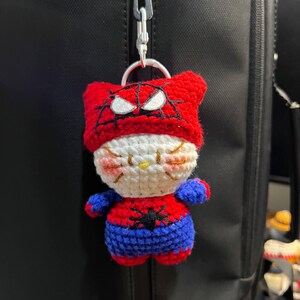 Puede incluir: Un llavero de peluche tejido a ganchillo con un personaje blanco similar a un gato, que lleva una capucha roja con un patr&oacute;n de telara&ntilde;a negra y parches oculares blancos. El personaje viste un traje rojo y azul con una ara&ntilde;a negra en el pecho.