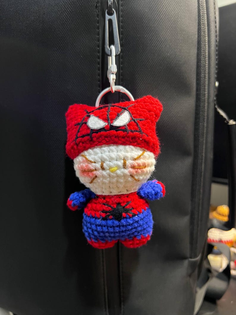 Puede incluir: Un llavero de peluche tejido a ganchillo con un personaje blanco similar a un gato, que lleva una capucha roja con un patr&oacute;n de telara&ntilde;a negra y parches oculares blancos. El personaje viste un traje rojo y azul con una ara&ntilde;a negra en el pecho.