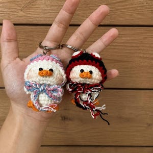 May include: Two handmade crochet duck keychains. One duck wears a pink and blue hat and bow tie, the other a black, red, and white hat and scarf. Both have orange beaks and feet, and are attached to silver keyrings.