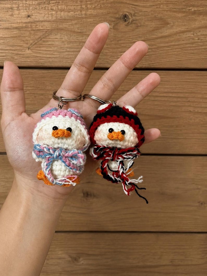 May include: Two handmade crochet duck keychains. One duck wears a pink and blue hat and bow tie, the other a black, red, and white hat and scarf. Both have orange beaks and feet, and are attached to silver keyrings.
