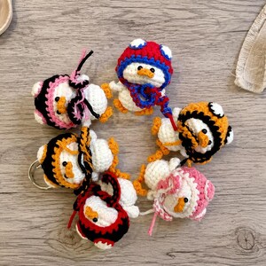 May include: A collection of handmade crochet duck keychains in various colors. Each duck has a white body, orange beak and feet, and is adorned with different colored hats and accessories, including red, black, pink, and orange. The keychains are arranged in a circular pattern.