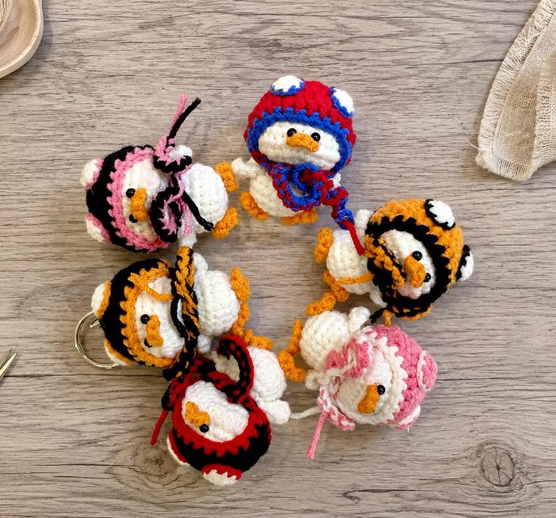 May include: A collection of handmade crochet duck keychains in various colors. Each duck has a white body, orange beak and feet, and is adorned with different colored hats and accessories, including red, black, pink, and orange. The keychains are arranged in a circular pattern.
