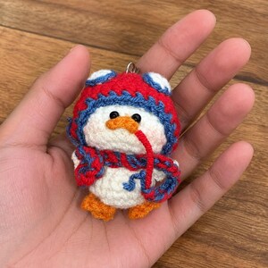 May include: A handmade crochet penguin keychain. The penguin is white with an orange beak and feet, black eyes, and a red and blue hat and scarf. The keychain is held in a hand.