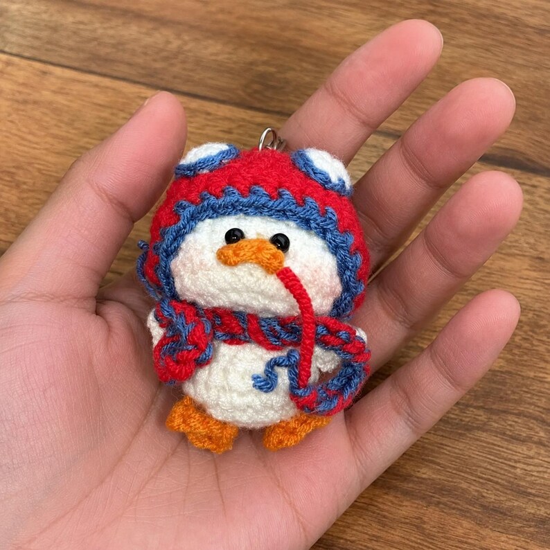 May include: A handmade crochet penguin keychain. The penguin is white with an orange beak and feet, black eyes, and a red and blue hat and scarf. The keychain is held in a hand.