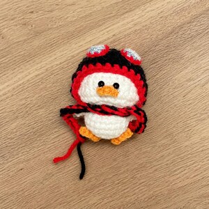 May include: A handmade crochet penguin with a white body, orange feet and beak, and a black, red, and silver hat and scarf. The penguin is a cute, whimsical design, perfect for a child's room or as a decorative item.