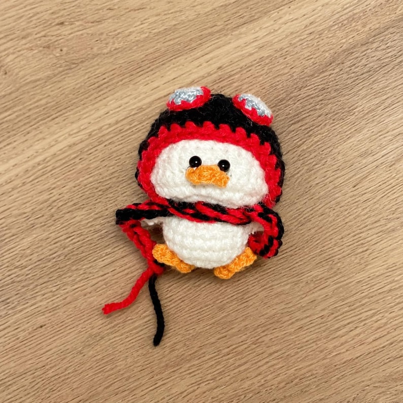 May include: A handmade crochet penguin with a white body, orange feet and beak, and a black, red, and silver hat and scarf. The penguin is a cute, whimsical design, perfect for a child's room or as a decorative item.