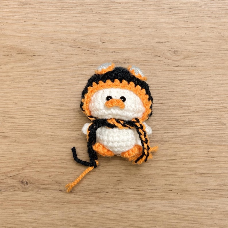 May include: A handmade crochet penguin doll. The penguin is white with orange feet and a matching orange beak. It wears a black and orange hat with silver accents and a black and orange scarf. The doll is approximately 4 inches tall.