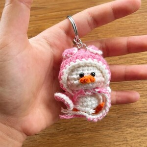 May include: A handmade crochet keychain featuring a small, white duck wearing a pink and white hooded outfit. The duck has an orange beak and black eyes. The keychain is attached to a silver metal ring.
