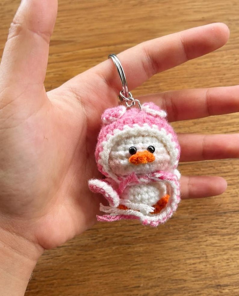 May include: A handmade crochet keychain featuring a small, white duck wearing a pink and white hooded outfit. The duck has an orange beak and black eyes. The keychain is attached to a silver metal ring.