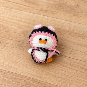 May include: A handmade crochet penguin doll with a black and pink hat and scarf. The penguin has a white body, orange beak, and black eyes. The doll is approximately 4 inches tall.