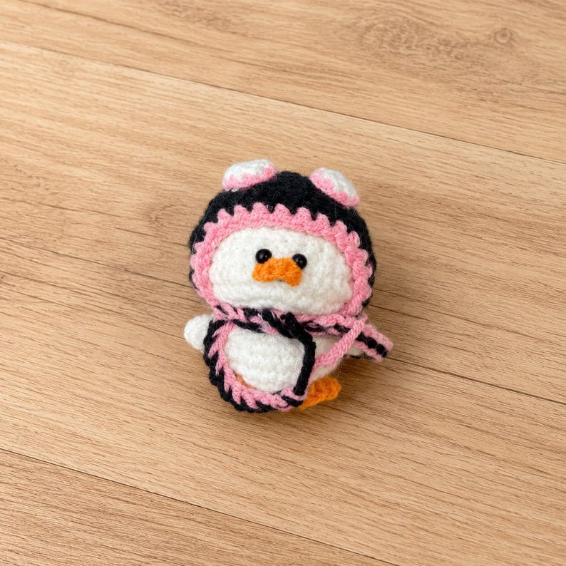 May include: A handmade crochet penguin doll with a black and pink hat and scarf. The penguin has a white body, orange beak, and black eyes. The doll is approximately 4 inches tall.