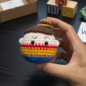 K&ouml;nnte beinhalten: Eine runde, geh&auml;kelte Amigurumi-Figur mit beigem Gesicht, braunem Haar in einem Dutt und schwarzen Perlenaugen. Der K&ouml;rper ist mit rotem, gelbem und blauem Garn gestreift. Die Figur hat kleine Ohren und wird vor einem dunklen Hintergrund gehalten.