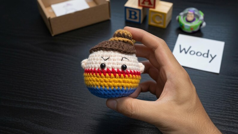 K&ouml;nnte beinhalten: Eine runde, geh&auml;kelte Amigurumi-Figur mit beigem Gesicht, braunem Haar in einem Dutt und schwarzen Perlenaugen. Der K&ouml;rper ist mit rotem, gelbem und blauem Garn gestreift. Die Figur hat kleine Ohren und wird vor einem dunklen Hintergrund gehalten.