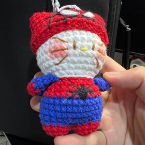 Puede incluir: Un llavero de peluche tejido a ganchillo con un personaje blanco de mejillas rosadas y bigotes, que viste un disfraz de superh&eacute;roe rojo y azul con un emblema de ara&ntilde;a negra en el pecho y una capucha roja con detalles de telara&ntilde;a negra.
