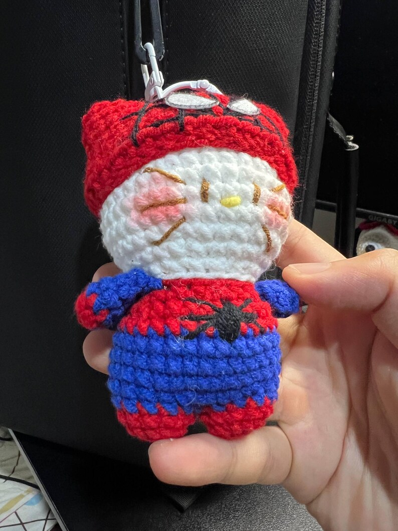 Puede incluir: Un llavero de peluche tejido a ganchillo con un personaje blanco de mejillas rosadas y bigotes, que viste un disfraz de superh&eacute;roe rojo y azul con un emblema de ara&ntilde;a negra en el pecho y una capucha roja con detalles de telara&ntilde;a negra.