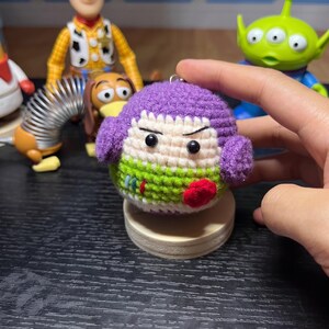 K&ouml;nnte beinhalten: Ein rundes, geh&auml;keltes Pl&uuml;schtier, inspiriert von einer Weltraum-Ranger-Figur, mit einer violetten Kapuze, einem cremefarbenen Gesicht mit schwarzen Perlenaugen und einem lindgr&uuml;nen K&ouml;rper mit roten und blauen Akzenten. Das Spielzeug hat eine Metall&ouml;se.