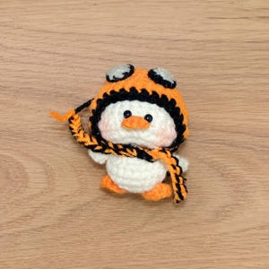 May include: A handmade crochet penguin wearing an orange and black aviator hat and scarf. The penguin has a white body, orange feet, and a small orange beak. The hat has black trim and goggles.
