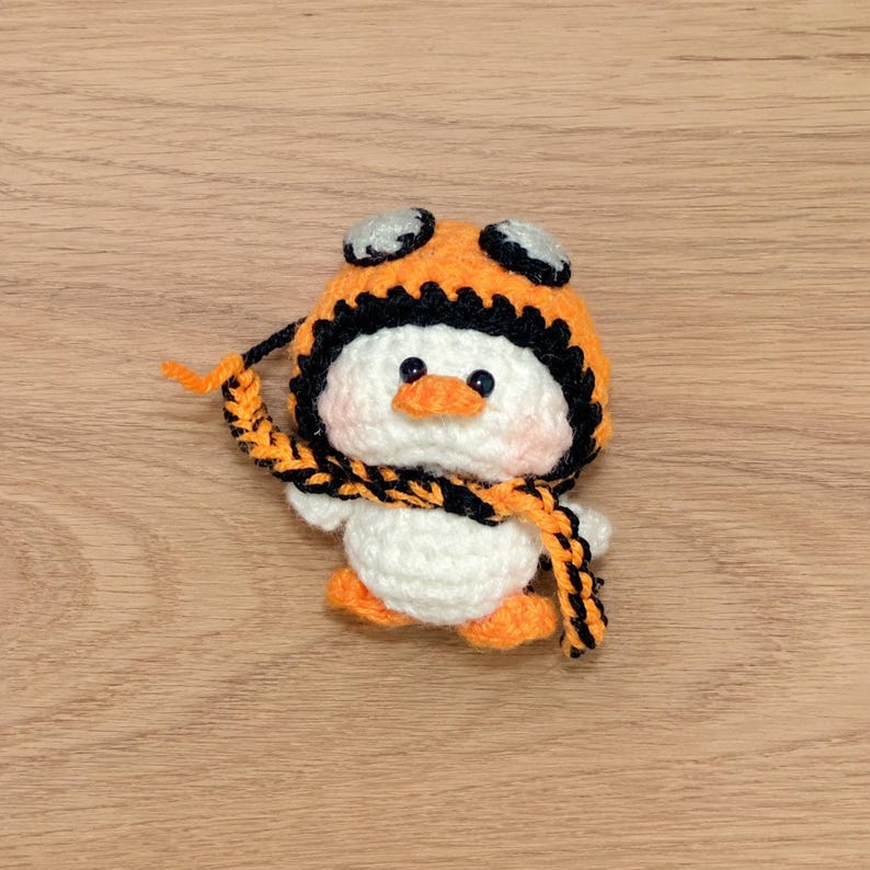 May include: A handmade crochet penguin wearing an orange and black aviator hat and scarf. The penguin has a white body, orange feet, and a small orange beak. The hat has black trim and goggles.