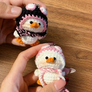 May include: Two handmade crochet duck keychains. One duck is wearing a black and pink hat and scarf, the other is wearing a pink and white hat and scarf. Both have orange beaks and feet, and black eyes. The ducks are white.