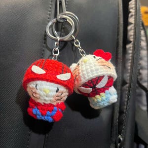 Puede incluir: Dos llaveros de ganchillo con personajes que llevan capuchas de superh&eacute;roe rojas y azules con parches oculares blancos. Un personaje tiene un lazo rojo, ambos tienen rasgos faciales bordados, bigotes y narices amarillas, en anillas plateadas.