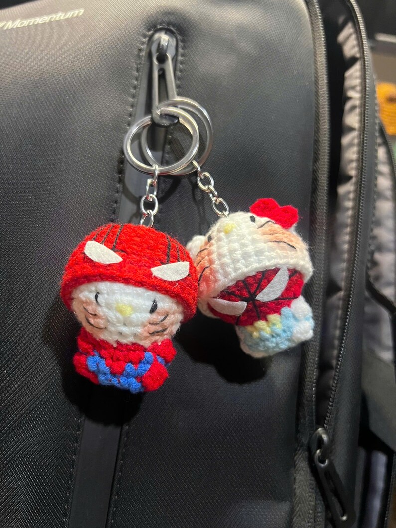 Puede incluir: Dos llaveros de ganchillo con personajes que llevan capuchas de superh&eacute;roe rojas y azules con parches oculares blancos. Un personaje tiene un lazo rojo, ambos tienen rasgos faciales bordados, bigotes y narices amarillas, en anillas plateadas.