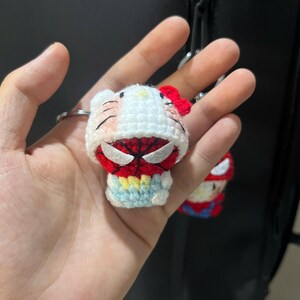 Puede incluir: Un peque&ntilde;o llavero de ganchillo hecho a mano que representa un personaje blanco con un lazo rojo, vestido con una capucha roja y negra con tem&aacute;tica de ara&ntilde;a. Tiene ojos blancos, cara roja con detalles de telara&ntilde;a negra y una base azul claro.