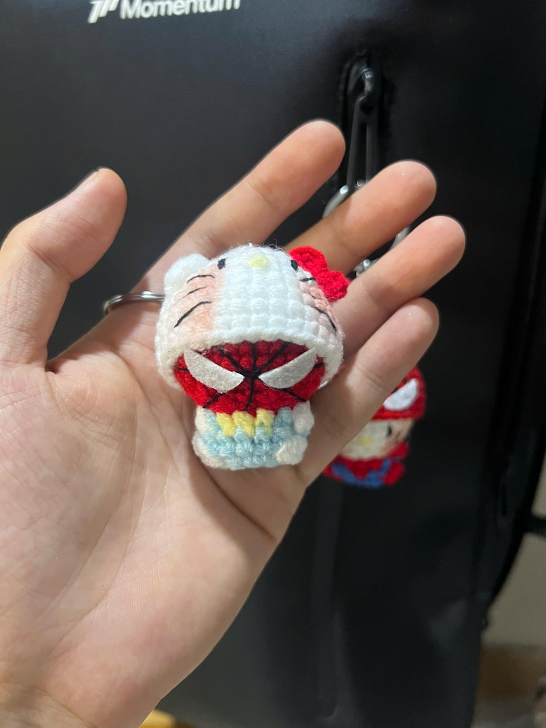 Puede incluir: Un peque&ntilde;o llavero de ganchillo hecho a mano que representa un personaje blanco con un lazo rojo, vestido con una capucha roja y negra con tem&aacute;tica de ara&ntilde;a. Tiene ojos blancos, cara roja con detalles de telara&ntilde;a negra y una base azul claro.