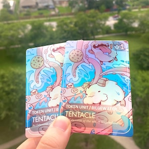 Peut inclure: Deux cartes de jeu rectangulaires avec une finition holographique présentant une illustration de dessin animé d'une créature blanche à cornes et d'un tentacule rose. Le texte indique TOKEN UNIT/BILGEWATER TENTACLE An Extension of the will of Nagakabouros.