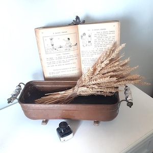 Peut inclure: Une jardinière rectangulaire en cuivre dotée de deux poignées avec des accents en céramique florale bleue et blanche et quatre petits pieds. Un bouquet de tiges de blé séchées repose à l'intérieur. Un livre ancien ouvert est placé derrière.