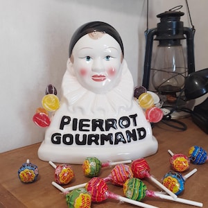 Op de afbeelding: Een wit keramieken Pierrot Gourmand displaybuste met een zwarte kap, geschilderde gelaatstrekken en een geplooide kraag. De basis is voorzien van de zwarte tekst PIERROT GOURMAND en houdt aan beide zijden diverse kleurrijke lolly's vast.