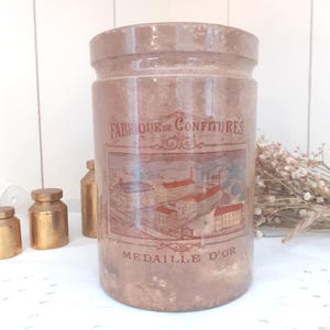 Peut inclure: Un pot cylindrique en céramique beige orné d'une illustration brune représentant une usine. Le texte indique FABRIQUE DE CONFITURES et MEDAILLE D'OR. Le pot présente une finition émaillée lisse avec un aspect vieilli et légèrement tacheté.