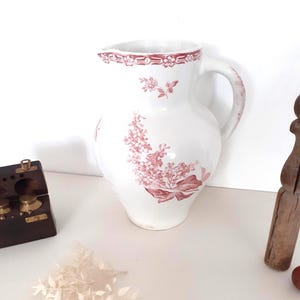 Pode incluir: Um jarro de cerâmica branca com um padrão floral decorativo em rosa. O design inclui uma borda detalhada ao redor da abertura, um grande ramo de flores no corpo e um padrão correspondente na alça. O jarro tem um bico e alça curvos.