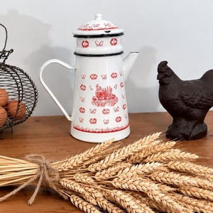 Könnte beinhalten: Eine weiße Emaille-Kaffeekanne mit einem roten Druckmuster aus Hühnern und Blumenmotiven, einem schwarzen Rand, einem passenden Deckel und einem geschwungenen Griff. Das Design zeigt mittig eine Henne, die in einem Nest sitzt.