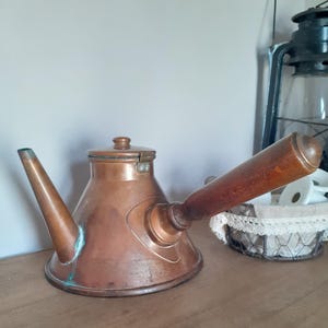May include: A copper kettle with a conical body, a long straight spout, and a thick wooden handle extending from the side. The kettle features a flat lid with a small knob and shows visible signs of oxidation with green patina near the base.