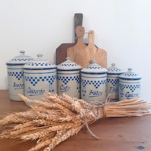 Könnte beinhalten: Ein Set aus sechs cremefarbenen Emaille-Vorratsdosen mit blauem Karomuster und blauen Beschriftungen: Sucre, Chicoree, Cafe, Pates, The und Epices. Jede Dose hat einen passenden Deckel und steht vor zwei hölzernen Schneidebrettern.