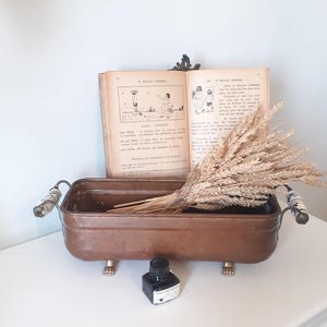 Peut inclure: Un bassin rectangulaire en cuivre doté de deux poignées latérales avec des inserts en céramique à motifs bleus et blancs, reposant sur quatre petits pieds métalliques. Un bouquet de blé séché est placé à l'intérieur devant un livre ancien ouvert.
