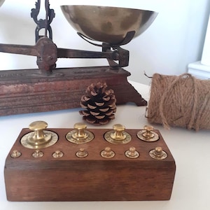 May include: A vintage cast iron balance scale with a brass bowl, accompanied by a rectangular wooden block holding a set of graduated brass weights. A small brown pinecone and a spool of natural jute twine are positioned near the scale.